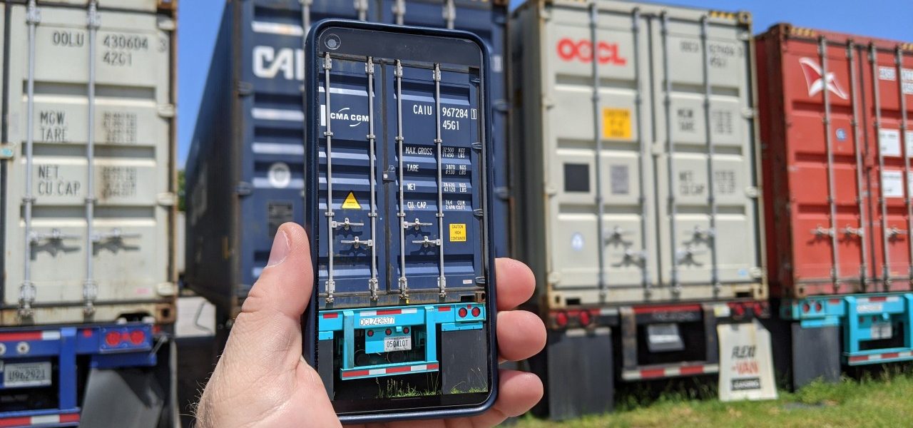 A person holding a smartphone that displays a digitized scan of a shipping container, standing in front of a row of stacked intermodal containers in a yard.