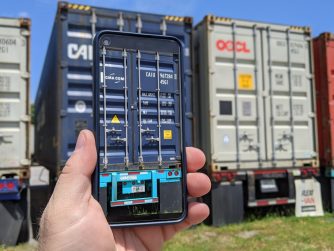 A person holding a smartphone that displays a digitized scan of a shipping container, standing in front of a row of stacked intermodal containers in a yard.