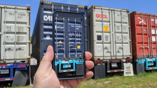 A person holding a smartphone that displays a digitized scan of a shipping container, standing in front of a row of stacked intermodal containers in a yard.