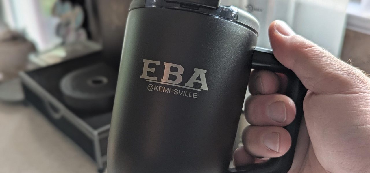 A hand holding a black insulated travel mug engraved with the EBA @Kempsville Entrepreneurship and Business Academy logo.