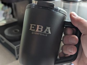 A hand holding a black insulated travel mug engraved with the EBA @Kempsville Entrepreneurship and Business Academy logo.
