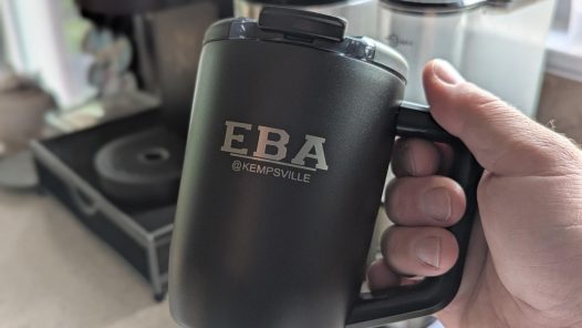 A hand holding a black insulated travel mug engraved with the EBA @Kempsville Entrepreneurship and Business Academy logo.