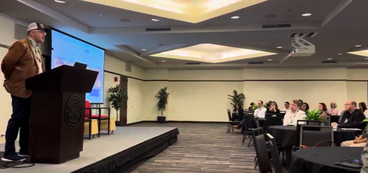 Chris Machut speaking at a podium in front of a seated audience while presenting at the ASCM San Diego event.