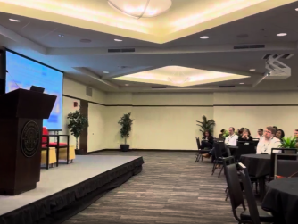 Chris Machut speaking at a podium in front of a seated audience while presenting at the ASCM San Diego event.