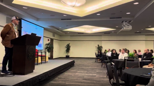Chris Machut speaking at a podium in front of a seated audience while presenting at the ASCM San Diego event.