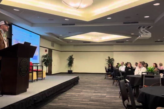 Chris Machut speaking at a podium in front of a seated audience while presenting at the ASCM San Diego event.